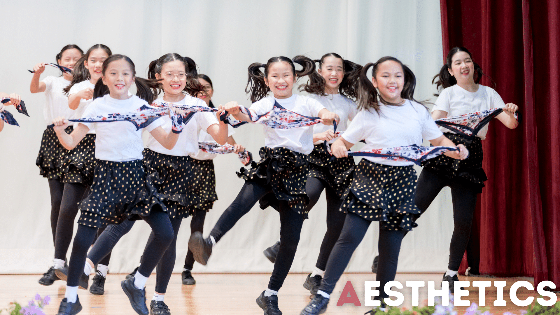 A group of young girls in white tops and polka dot skirts dance on stage holding floral scarves.